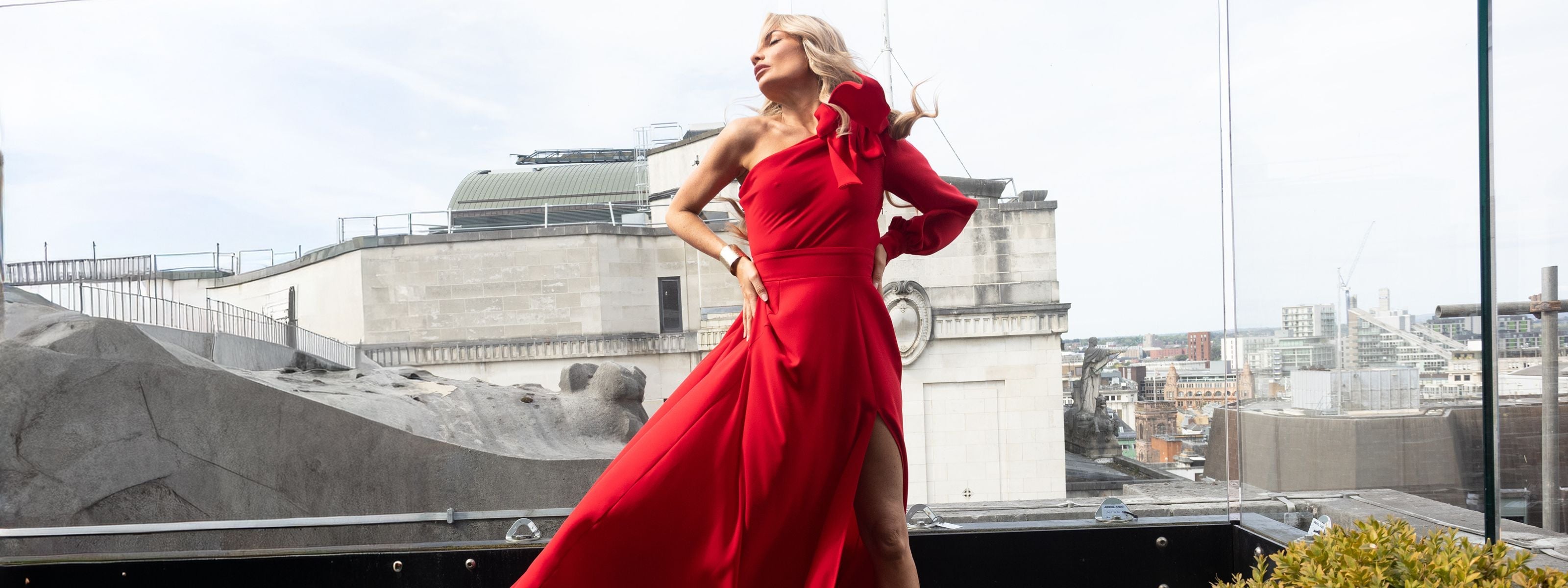 Model wearing a dramatic red one-shoulder gown by RHOEN, designed by Rebecca Rhoades, featuring a statement bow detail, long sleeve, and thigh-high slit, photographed on a city rooftop with flowing fabric creating a bold and elegant silhouette.